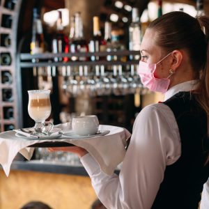 A female Waiter of European appearance in a medical mask serves Latte coffee